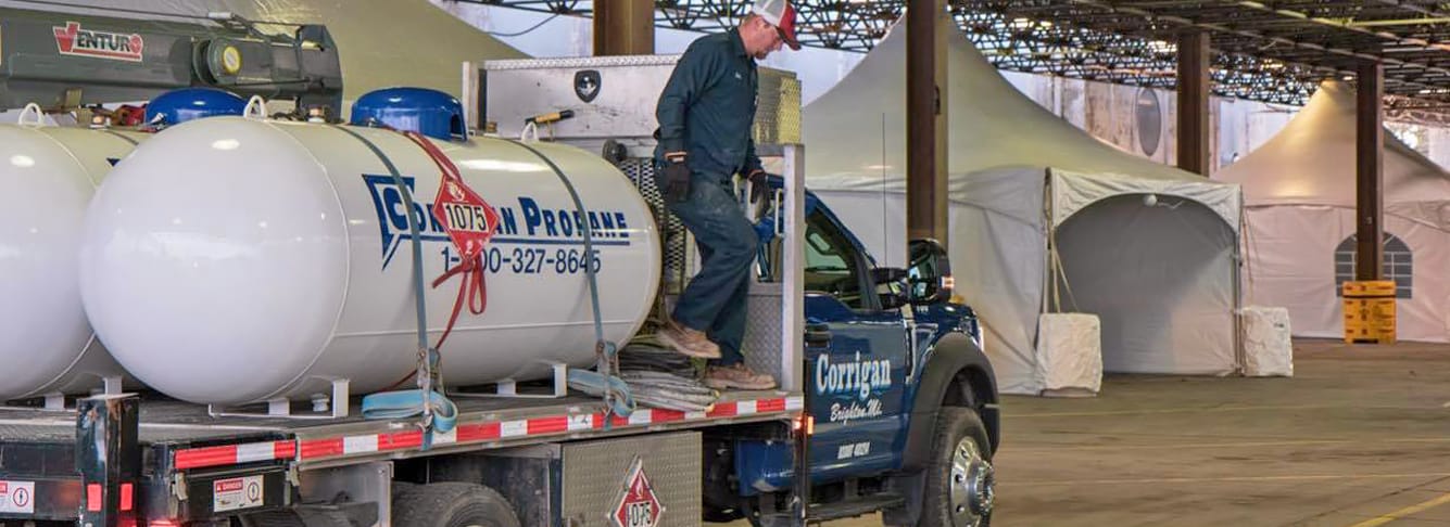 A worker in protective clothing steps off a Corrigan-branded propane delivery truck loaded with large white propane tanks inside a spacious industrial building with tented structures in the background.